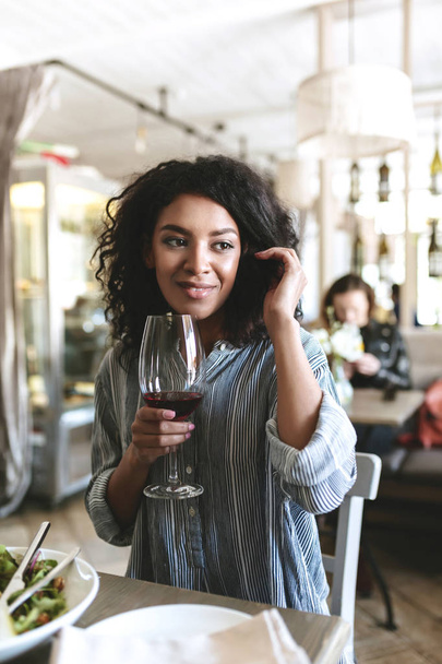 Jeune fille afro-américaine assise au restaurant avec un verre de vin rouge à la main. Portrait de belle fille aux cheveux bouclés foncés qui regarde de côté et rangeant ses cheveux au café
 - Photo, image