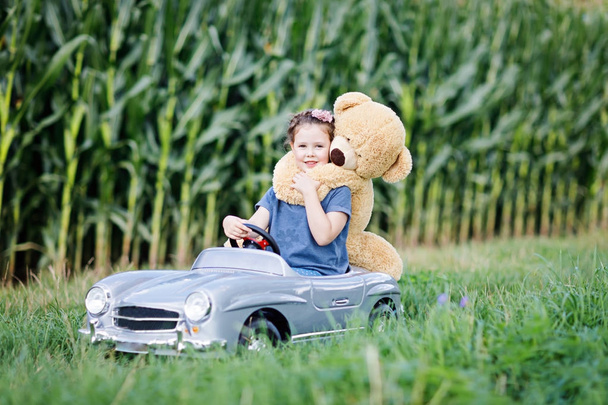 Маленькая дошкольница водит большую игрушечную машину и веселится, играя с большим плюшевым игрушечным медведем
 - Фото, изображение