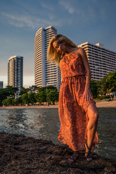 Beautiful blonde in a light red dress stands by the sea on the backdrop luxurious resort in the late afternoon sunlight. Posing for the camera - Foto, afbeelding