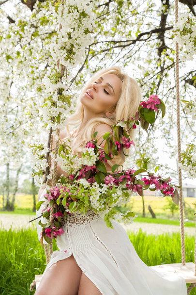 春の花束を保持スプリング ドレスを着て木のブランコに乗って見事なブロンドのセクシーな自然女性スイング - 写真・画像
