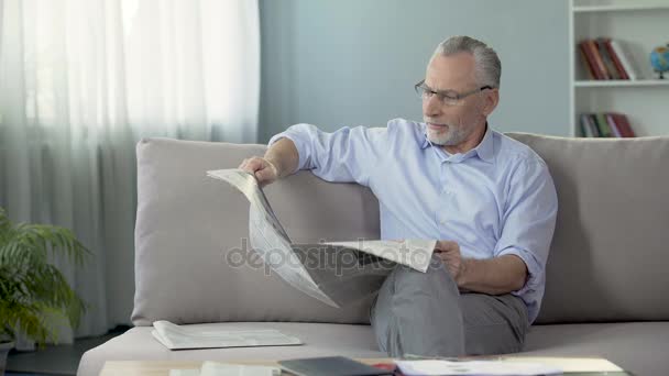 Senior male sitting on couch and reading newspaper, morning ritual, press - Felvétel, videó