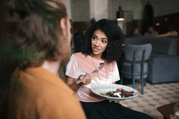 Улыбающаяся афроамериканка сидит в кафе с тарелкой салата в руке. Молодая девушка с темными кудрявыми волосами разговаривает с другом
 - Фото, изображение