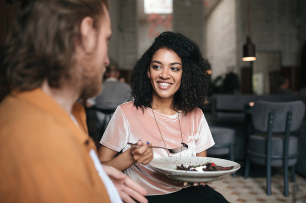 Lachende meisje zittend in restaurant met vriend. Mooie Afro-Amerikaanse dame zit op café met salade in de hand - Foto, afbeelding