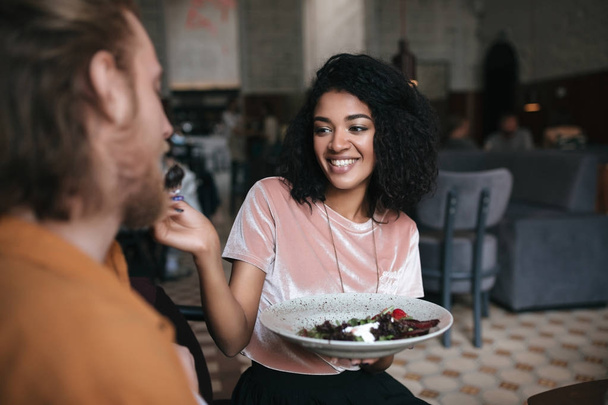 Радостная девушка сидит в ресторане и разговаривает с другом. Улыбающаяся афроамериканка сидит в кафе с тарелкой салата в руке
 - Фото, изображение