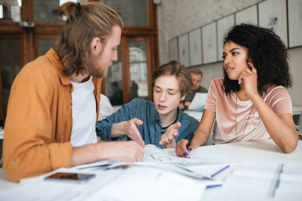 Portrait of young people emotionally discussing something in office. Two boys with blond hair and girl with dark curly hair sitting and working together in classroom - Photo, Image