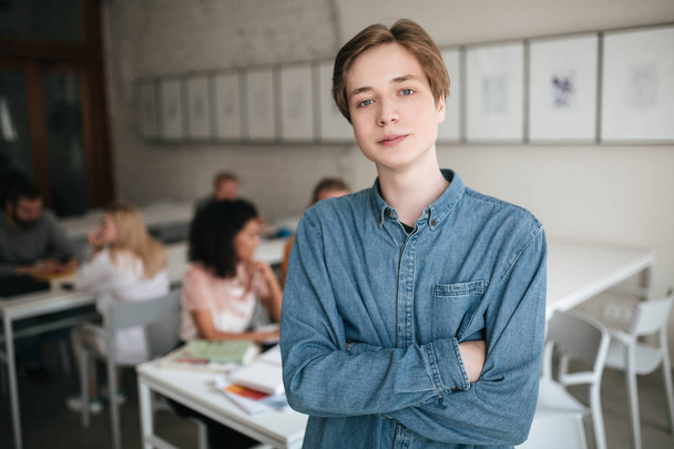 Cool boy in denim shirt standing with clasped hands in classroom with students on background - Fotoğraf, Görsel