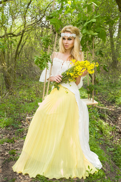 atemberaubende blonde sexy natürliche Frau Schaukel auf einer Schaukel auf einem Baum trägt ein Frühlingskleid hält einen Strauß Frühling - Foto, Bild