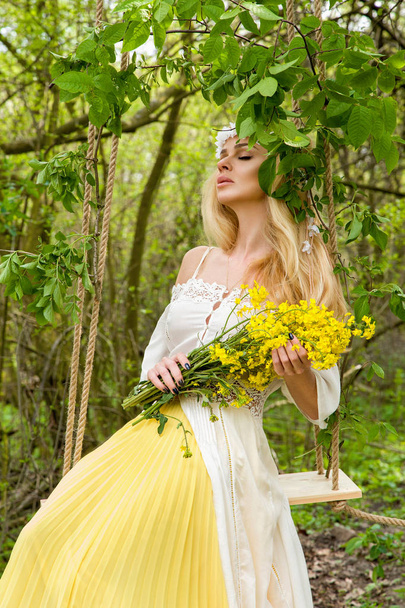 Приголомшлива блондинка сексуальна натуральна жінка гойдається на дереві в весняній сукні, що тримає букет весни
 - Фото, зображення