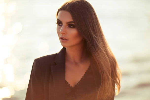 Portrait of pretty serious business woman is wearing black official clothes near wooden wall over sunset lights and sea - Photo, Image