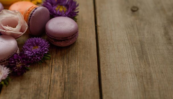 violette und gelbe Macarons und Blumen auf hölzernem Tischhintergrund. buntes französisches Dessert mit frischen Blumen - Foto, Bild