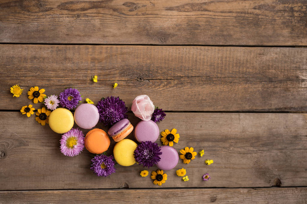 Macarons και λουλούδια στο φόντο του ένα ξύλινο τραπέζι. Πολύχρωμο γαλλικό επιδόρπιο με φρέσκα λουλούδια. Διαβάθμιση μεταξύ μοβ και κίτρινο. Θέση για το κείμενο - Φωτογραφία, εικόνα