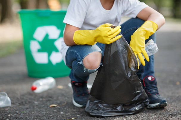 Hands in yellow gloves picking up empty of bottle plastic into bin bag on asphalt, volunteer and purity concept. Enviromental protection. - 写真・画像
