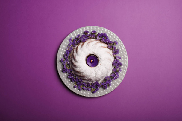 A plate with little purple flowers, a ring cake covered with powdered sugar, a piping hot bakery product for celebrations on a bright violet background, top view. - Фото, изображение