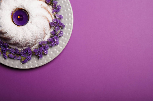 A ring cake covered with powdered sugar on a plate decorated with little purple flowers and a lit candle on a bright violet background, copy space, top view. - Фото, изображение