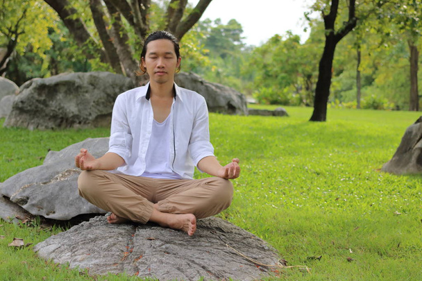 Hombre yogui joven haciendo meditación de yoga mientras está sentado en posición de loto sobre la roca en el parque
. - Foto, Imagen