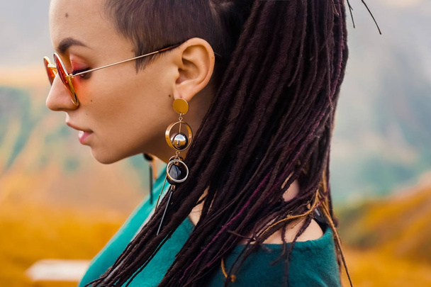 portrait of a young beautiful hipster girl in sunglasses with hair dreadlock in a boho gypsy hippie style clothes on a background of cloudy sky and autumn mountains of Georgia dancing - Photo, Image