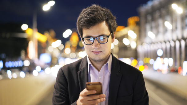 Handsome young businessman in glasses web surfing and smiling, smartphone, night - Footage, Video