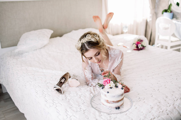 Portrait of a young charming blonde in white lingerie. On a large bed with cake and shoes - Photo, image