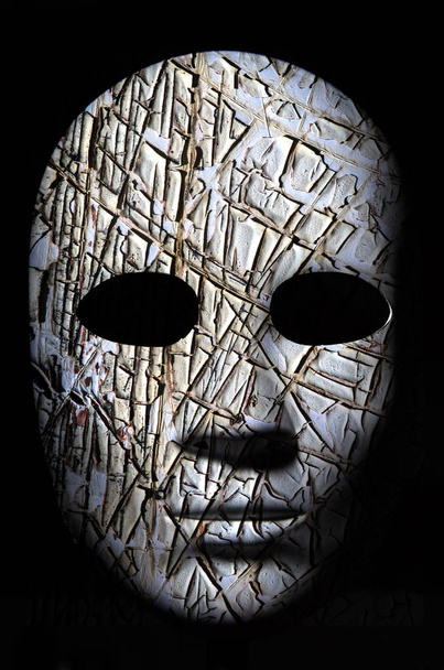 Máscara texturizada con superficie pintada de madera agrietada, expresión neutra sobre fondo oscuro
. - Foto, Imagen