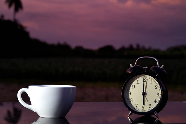 Bahçedeki ahşap masa üzerinde siyah çan saat ve fincan sıcak kahve  - Fotoğraf, Görsel