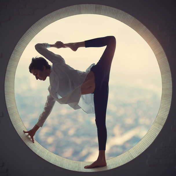Beautiful sporty fit yogi woman practices yoga asana Natarajasana - Lord Of The Dance pose in a round window at sunset - Foto, afbeelding