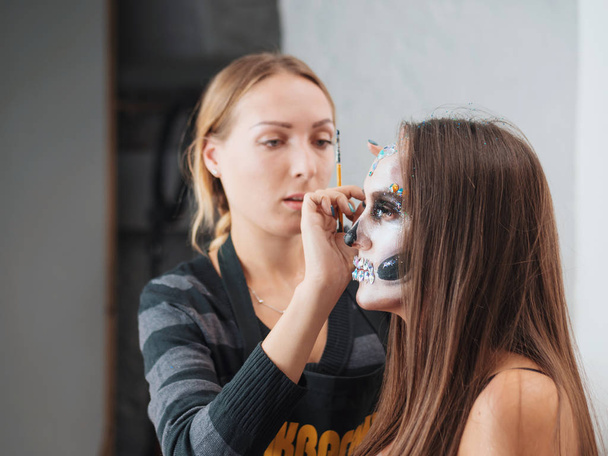 Make-up artist make the girl halloween make up in studio.Halloween face art.Woman applies on professional greasepaint on the face of girl.War-paint with blood, scars and wounds. - Photo, Image