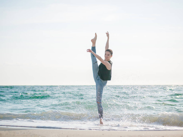 Beautiful scene of a dancing ballerina in black top and jeans on sandy beach ocean or sea in morning. Beautiful brunette woman practicing stretching and exercises.Copy space - Foto, immagini