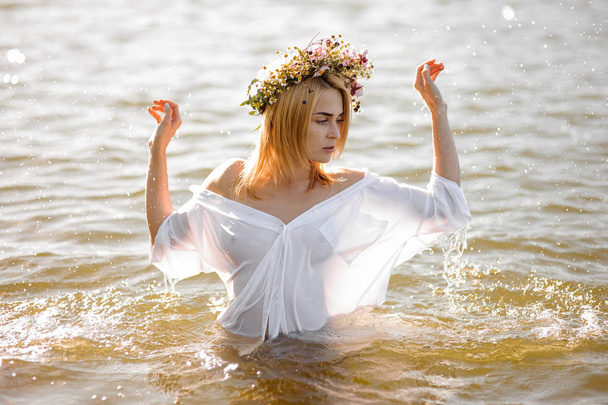 Girl in a white shirt and floral wreath standing in water - Fotó, kép