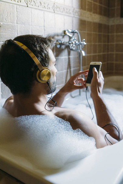 naked guy in bathroom with smartphone and headphones - Foto, Imagem