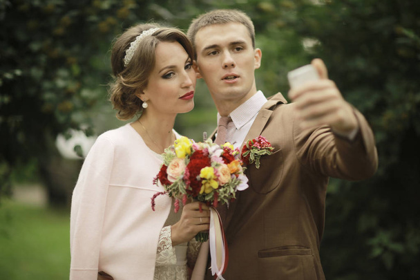 beautiful young bride and handsome groom posing in summer park on wedding day and taking selfie  - Zdjęcie, obraz