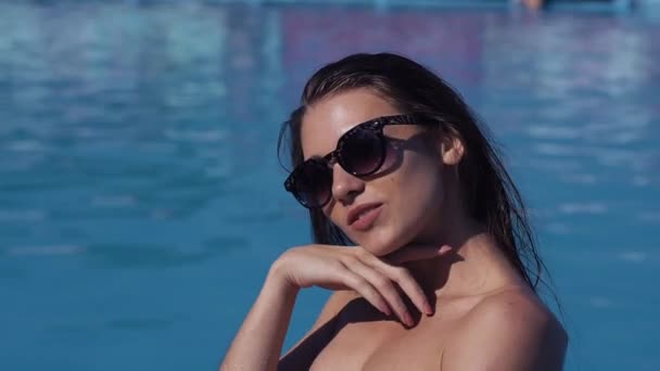 Close up young beautiful girl in sunglasses in swim wear near the swimming pool looking at the camera - Felvétel, videó