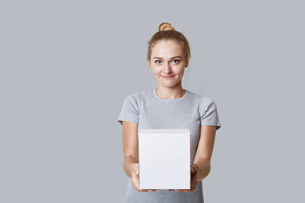 Young beautiful blonde female with hair bun holds white box in hands, with copy space for your text, isolated over grey background. Attractive woman demonstrates box, has appealing appearance - Foto, Imagen