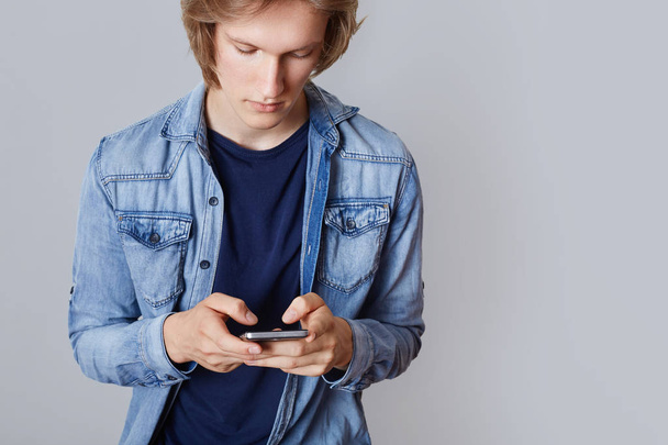Cropped shot of male teenager in denim shirt, holds modern smart phone, plays games online, surf social networks and writes posts, being internet addicted. Hipster guy communicates with friends - Фото, зображення