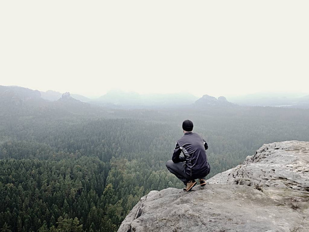 Vista trasera de excursionista solo en traje oscuro al aire libre sentarse en el borde. Pico rocoso agudo sobre el valle de las montañas
 - Foto, Imagen