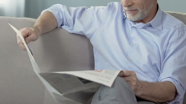 Bearded senior man sitting on couch and reading newspaper, weekend morning news - Video, Çekim