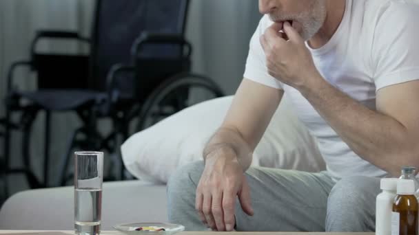 Elderly male taking pill, looking at wheelchair standing at background, trauma - Imágenes, Vídeo