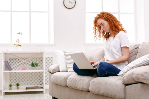 Young redhead girl with laptop sitting on beige couch - Фото, изображение