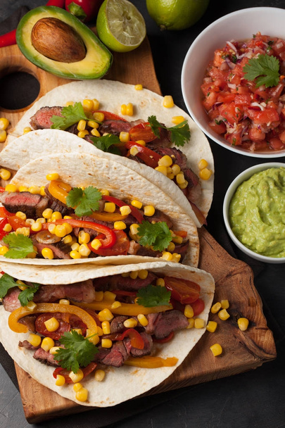 Three Mexican tacos with marbled beef, black Angus and vegetables on wooden Board on a dark stone background. Mexican dish with sauces guacamole and salsa in bowls. Top view - Photo, Image