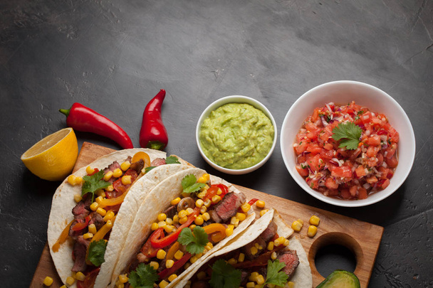 Trzy meksykańskie tacos z wołowiną marmurkowym, black Angus i warzywa na desce na ciemnym tle kamienia. Meksykańskie danie z guacamole sosy i salsy w miskach. Widok z góry z miejsca kopii - Zdjęcie, obraz