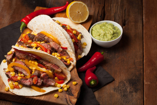 Three Mexican tacos with marbled beef, black Angus and vegetables on old rustic table. Mexican dish with sauces guacamole and salsa in bowls - Foto, imagen