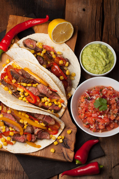 Three Mexican tacos with marbled beef, black Angus and vegetables on old rustic table. Mexican dish with sauces guacamole and salsa in bowls. top view - Photo, Image