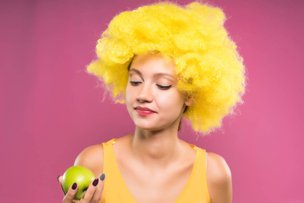 cute girl in a bright yellow wig is holding a green apple in her hands and looking at it, isolated on a pink background - Fotó, kép