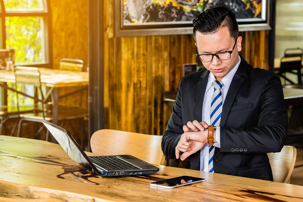Азиатский бизнесмен смотрит наручные часы во время работы со смартфоном и ноутбуком
. - Фото, изображение