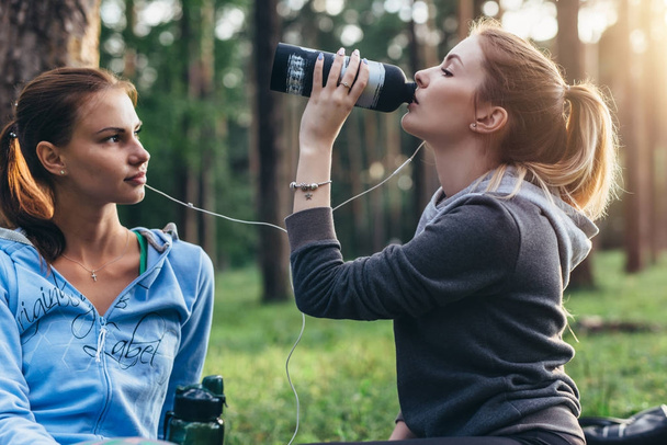Kaksi naisurheilijaa puhuu taukoa treenattuaan juomavettä puistossa.
 - Valokuva, kuva