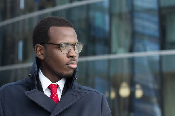 Outdoor portrait of handsome African businessman pictured in urban environment dressed in coat with lifted collar looking to street through eyeglasses with expression of serious concentration on work - Foto, Imagem