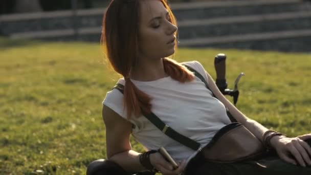 Redhaired woman cyclist resting on green lawn in summer park - Materiał filmowy, wideo