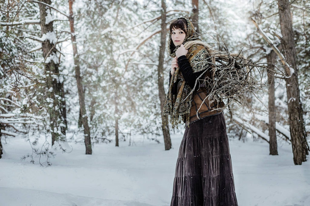 Una joven vestida con ropa vieja está de pie con broza en un bosque de invierno. personaje del cuento de hadas "doce meses
" - Foto, Imagen