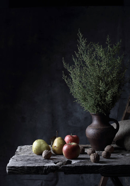 Stunning still life with walnuts, pears, apples and jug with green plants in it, on wooden table in dark studio - Photo, Image