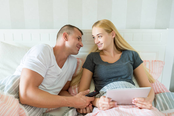 Young attractive caucasian couple in bed. Woman holding digital tablet, man lovingly looking at her, hoping to catch her attention. Both with cheeky smile. - Photo, image