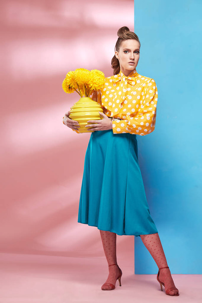 Beautiful young sexy woman model wearing yellow blouse with white polka-dot, blue skirt and pink tights in pin-up style, holding vase with yellow flowers and posing in studio with blue and pink background  - 写真・画像